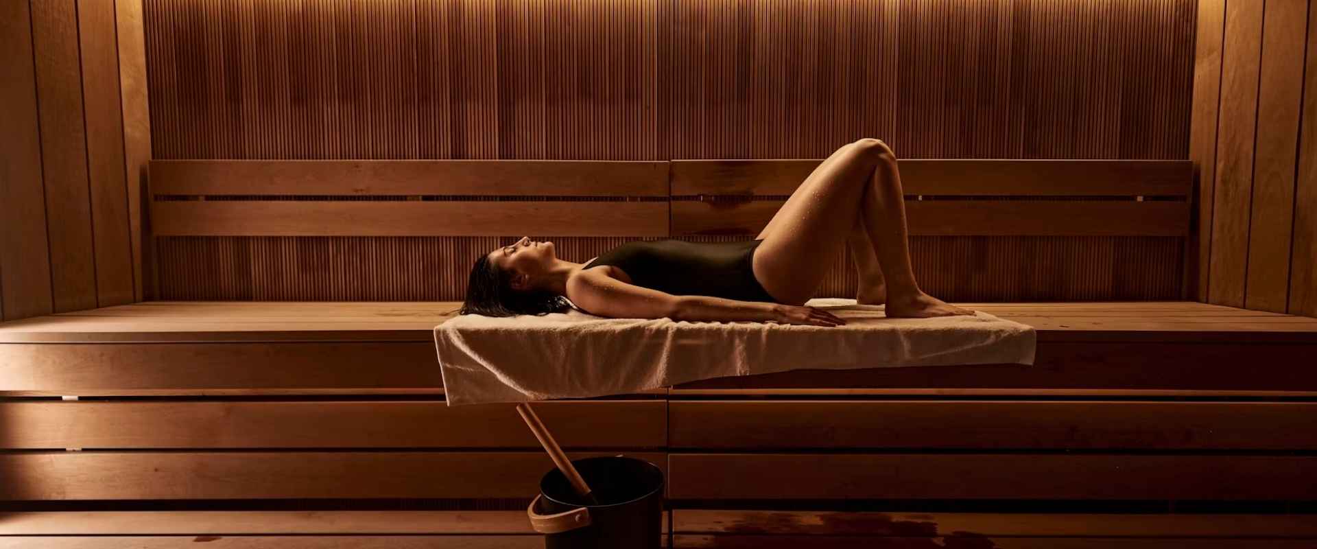 A woman in a black swimsuit lies on a wooden sauna bench with a white towel draped over it, inside a wooden sauna room with warm ambient lighting.
