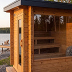 A wooden outdoor sauna cabin with a glass door, interior bench, and large window overlooking a lakeside deck at sunset.