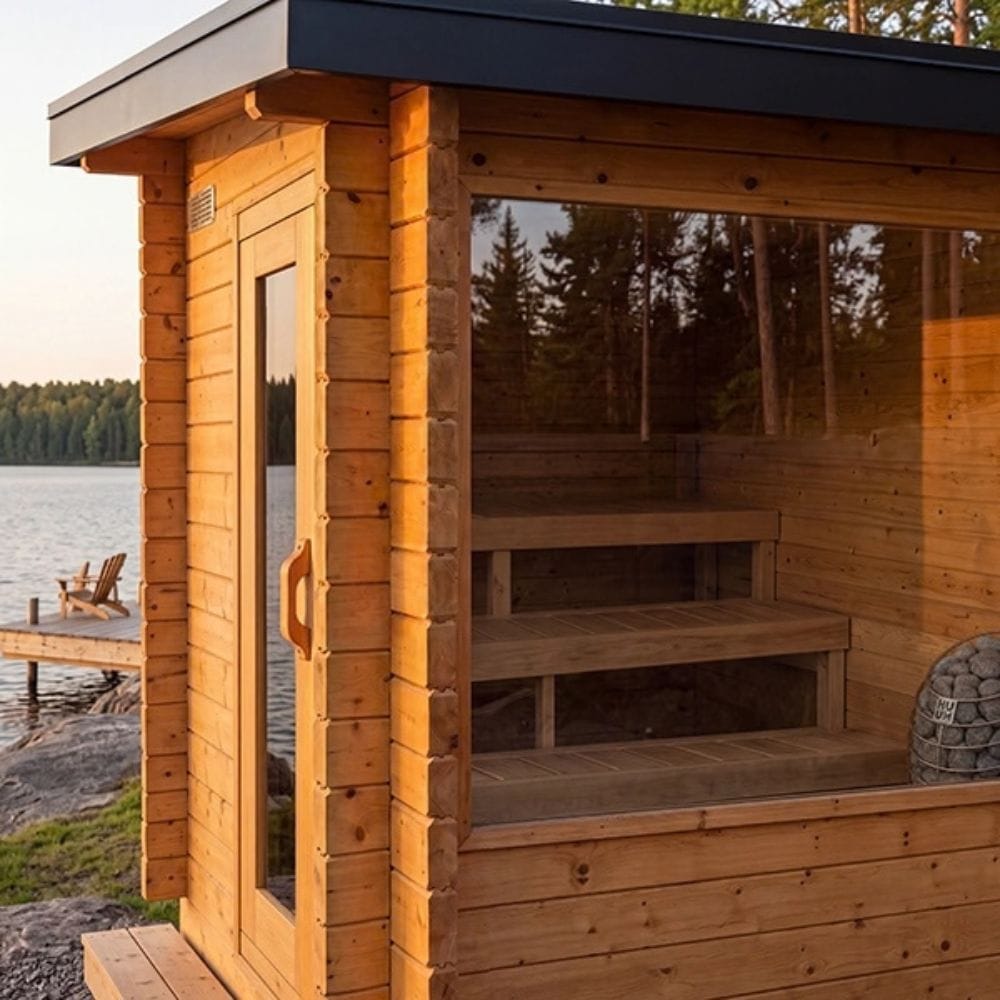 A wooden outdoor sauna cabin with a glass door, interior bench, and large window overlooking a lakeside deck at sunset.