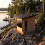 A small wooden outdoor sauna cabin with a dark metal roof sits on rocky shoreline beside a lake, with trees and a dock in the background.
