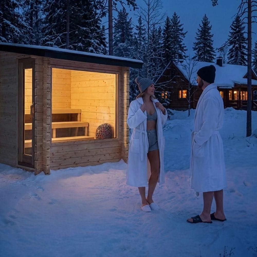 Two people in white robes stand in a snowy outdoor setting beside a small wooden sauna cabin with warm yellow light glowing from inside, with snow-covered trees and cabins in the background at dusk.
