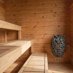 A wooden sauna interior with two wooden benches, wood-paneled walls, and a hanging metal basket filled with dark heating stones.