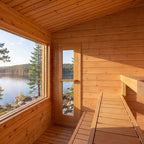 A wooden cabin interior with a large window overlooking a lake and forest, featuring a glass door and built-in wooden benches.