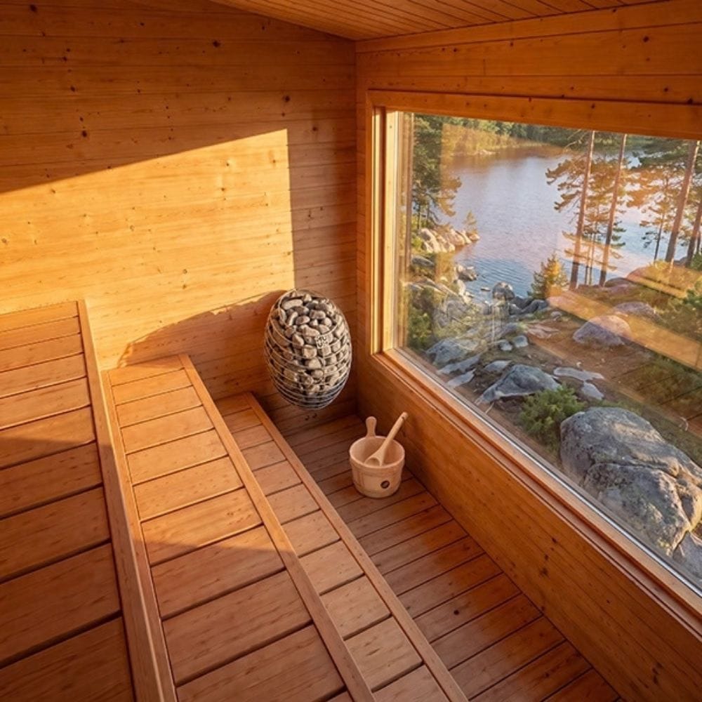 A wooden sauna interior with a large window overlooking a lake and forest, featuring a wood bench, hanging stone basket, and a small wooden bowl with a spoon.