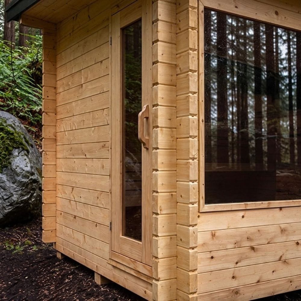 A close-up of a light natural wood cabin exterior, showing a glass door with a wooden handle and a large window reflecting a forest background.