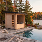 A small light wood cabin with a glass door and large window sits beside a stone pool, surrounded by trees and a lake at sunset.
