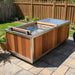 A rectangular outdoor hot tub or plunge pool with a wood-look exterior and stainless steel trim, shown on a stone patio in a backyard with a wooden fence and grass.