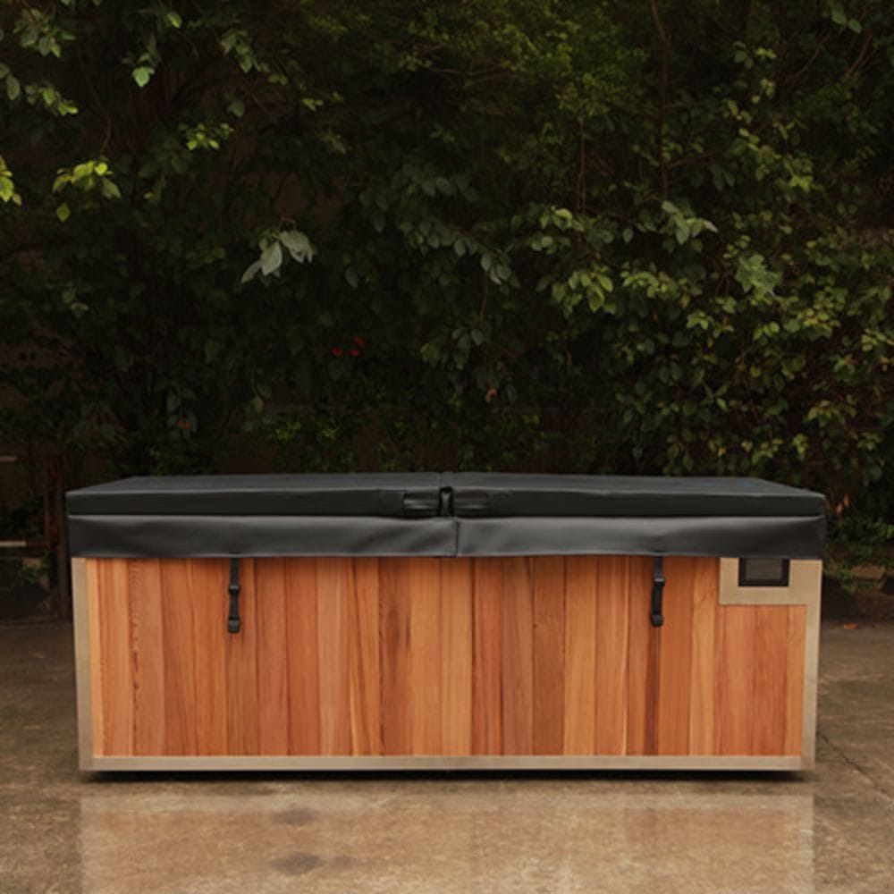 A rectangular outdoor hot tub or spa with a wood-look brown front panel, light beige trim, and a dark gray cover, photographed on a patio with dense green foliage in the background.