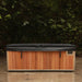 A rectangular outdoor hot tub or spa with a wood-look brown front panel, light beige trim, and a dark gray cover, photographed on a patio with dense green foliage in the background.