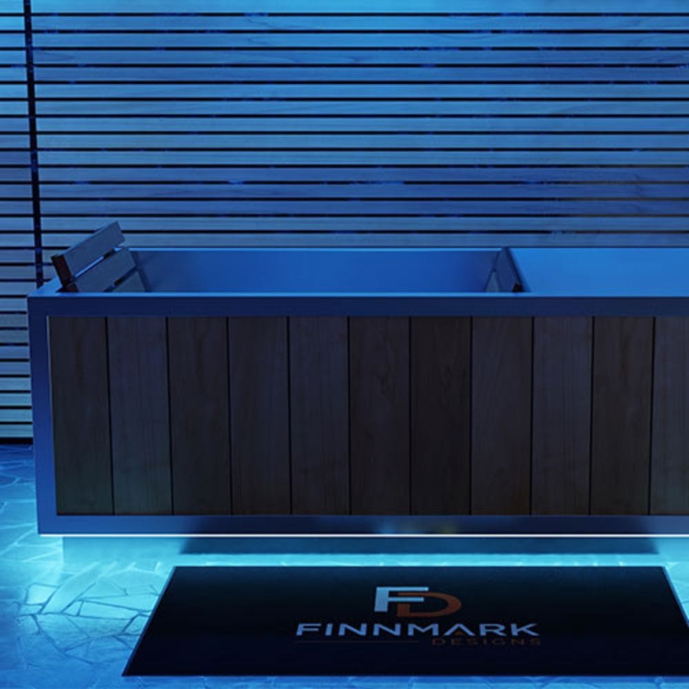 A modern rectangular hot tub or spa with a dark wood-paneled front and light blue interior, shown in a blue-lit bathroom setting with a black FINMARK DESIGNS floor mat in front.