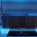 A modern rectangular hot tub or spa with a dark wood-paneled front and light blue interior, shown in a blue-lit bathroom setting with a black FINMARK DESIGNS floor mat in front.