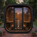 A modern outdoor sauna cabin with a rounded rectangular shape, featuring a black metal frame, large glass doors and side windows, wooden walls and benches, and a stone-burning stove inside, set on a brick patio in a landscaped garden at dusk.