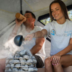A couple relaxing inside a sauna tent, with a woman pouring hot water from a ladle into a metal steaming stone basket. The woman wears a light gray t-shirt with the blue and teal 'saunalife' logo, and the man holds a wooden mug.