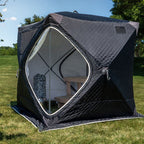 A black diamond-quilted outdoor shower tent pitched on a green grass lawn under a blue sky with trees in the background. The tent features a large hexagonal opening with a white zipper, showing a wooden bench and a stack of gray stone tiles inside.
