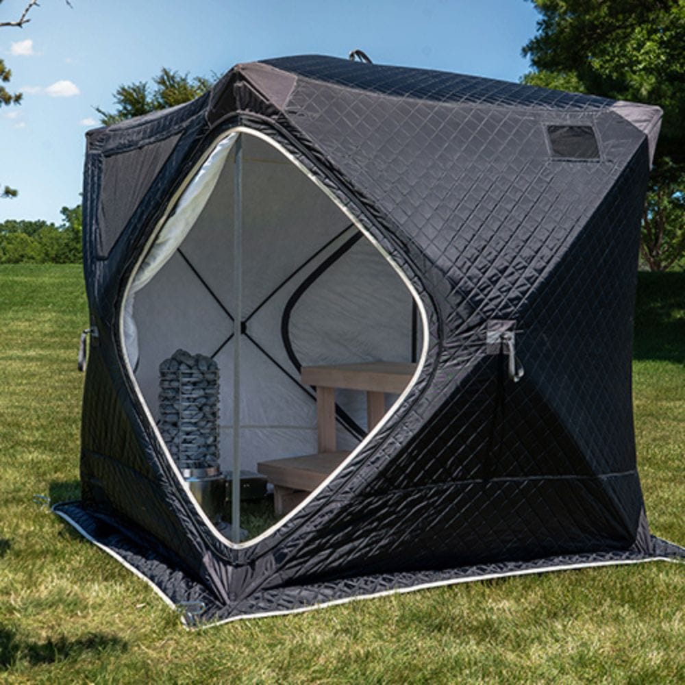 A black diamond-quilted outdoor shower tent pitched on a green grass lawn under a blue sky with trees in the background. The tent features a large hexagonal opening with a white zipper, showing a wooden bench and a stack of gray stone tiles inside.