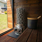 A stainless steel stone heater with a wire mesh cylinder filled with gray stones sits on a wooden deck beside a window, next to a dark gray bucket with a bamboo lid.