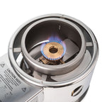 Close-up of a compact stainless steel gas stove burner ignited with a blue and orange flame, showing the burner head, inner ring, outer pot, and part of the stove body with printed caution labels.