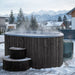 Wooden hot tub with steam rising in a snowy outdoor setting with mountains in the background.