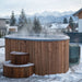 Wooden hot tub with steam rising in a snowy outdoor setting with mountains in the background.