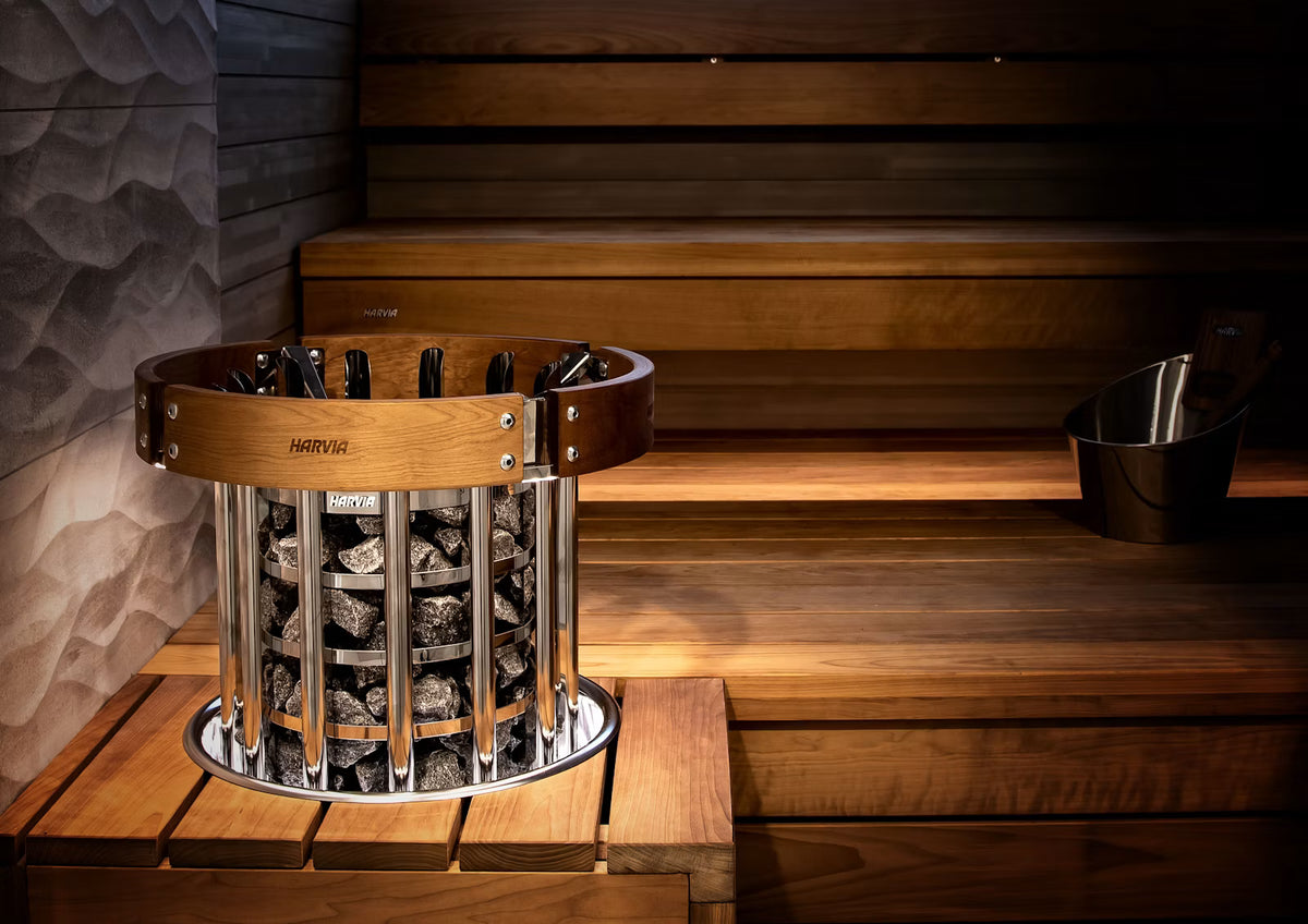 A cylindrical sauna fire basket filled with dark wood logs, set on a wooden bench inside a sauna with wood steps and a metal bucket nearby.